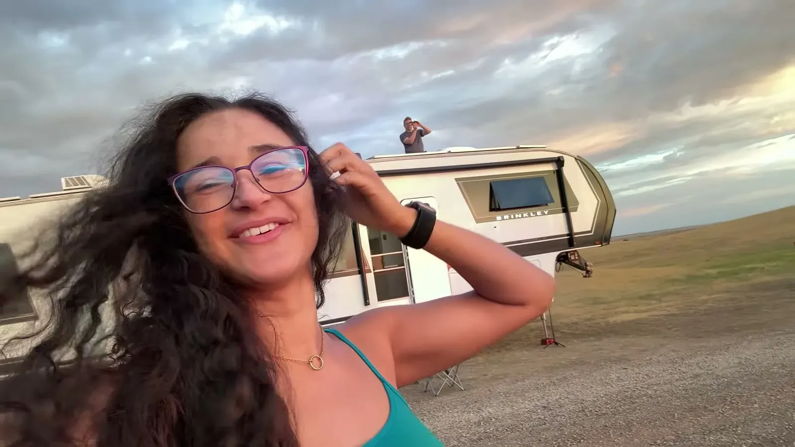 Fabiola in the foreground while Nathan stands on the fifth wheel roof checking the solar panels under storm clouds