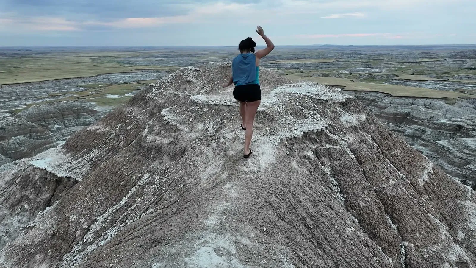 Fabiola with an arm raised walking a narrow Badlands ridge line above the prairie near our free campsite