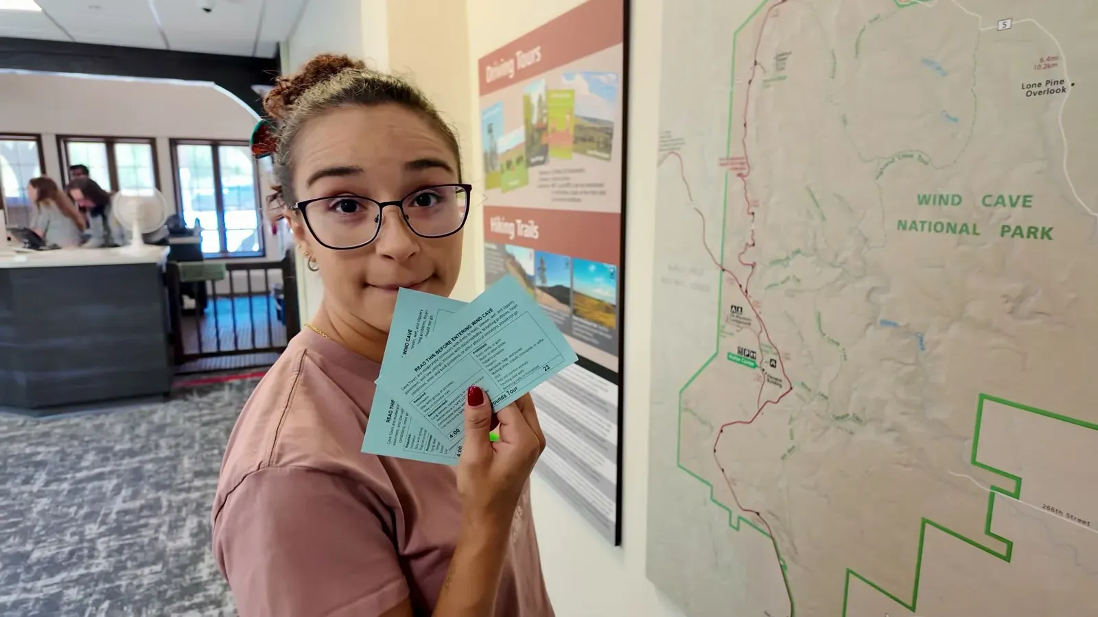 Fabiola holding cave tour tickets beside a large Wind Cave National Park trail map in the visitor center