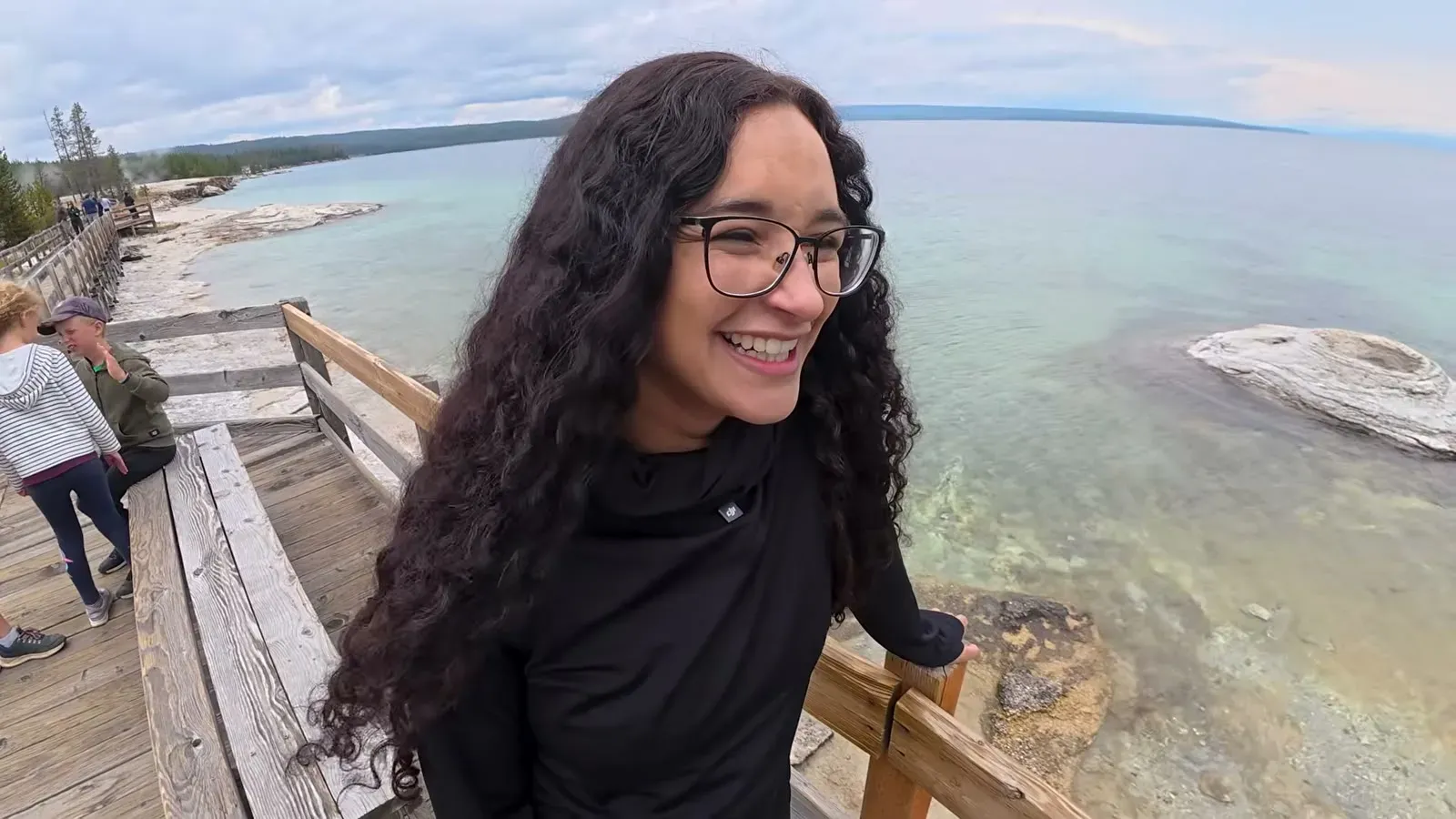 Fabiola smiling on the West Thumb boardwalk with Yellowstone Lake and a white geyser cone in the shallow water behind her