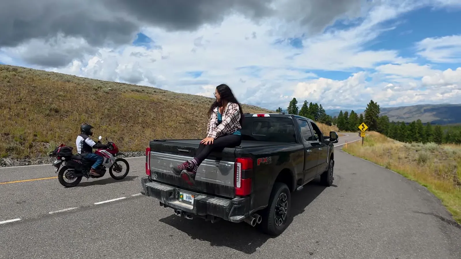 Fabiola sitting on the tailgate of our black pickup at a mountain pullout in Yellowstone as a motorcycle rides past