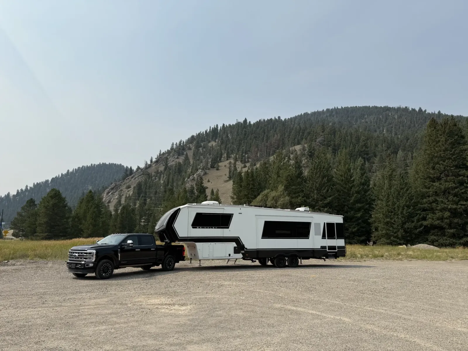 Our black Ford F-350 hitched to our Brinkley fifth wheel on a gooseneck ball, parked in a gravel lot below a forested hill