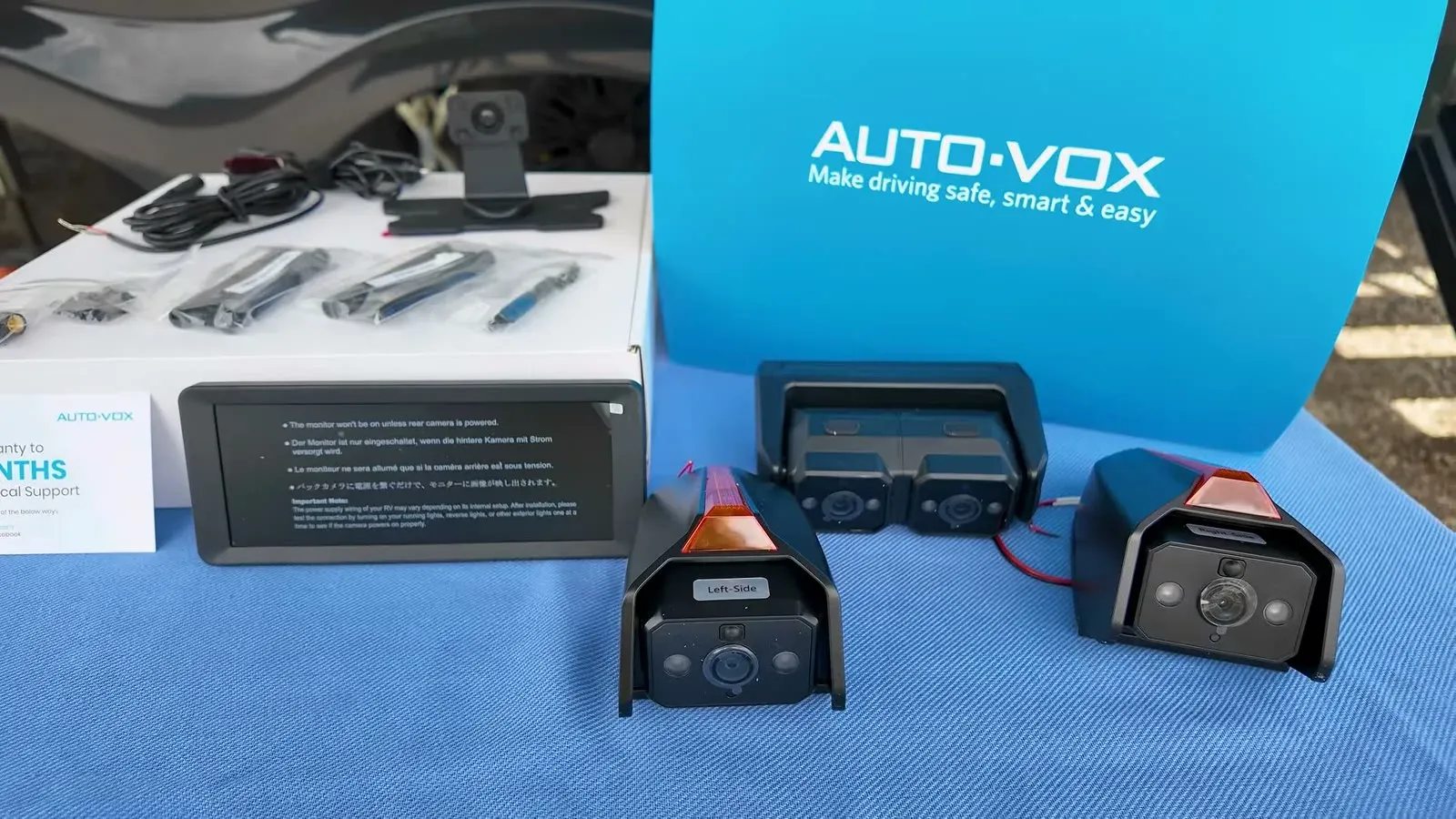 The AUTO-VOX CampSync kit laid out on a blue tablecloth: the monitor, the two-camera rear unit and the two side cameras with amber lights