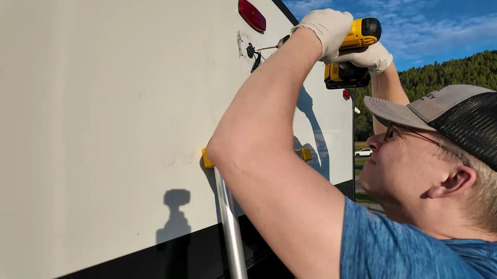 Nathan in work gloves on a ladder, drilling new mounting holes for the rear camera on the back wall of our fifth wheel