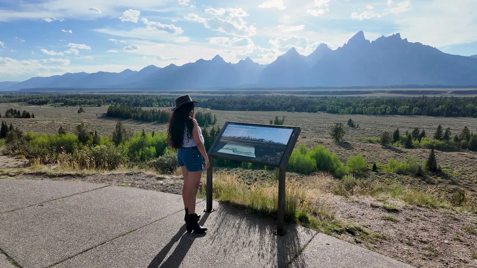 Fabiola beside an interpretive sign at Teton Point Turnout, with the valley and the Teton Range beyond
