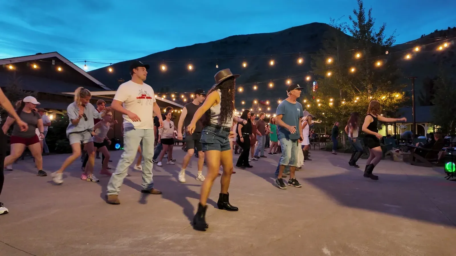 A crowd line dancing outdoors under string lights at dusk in Jackson, Wyoming