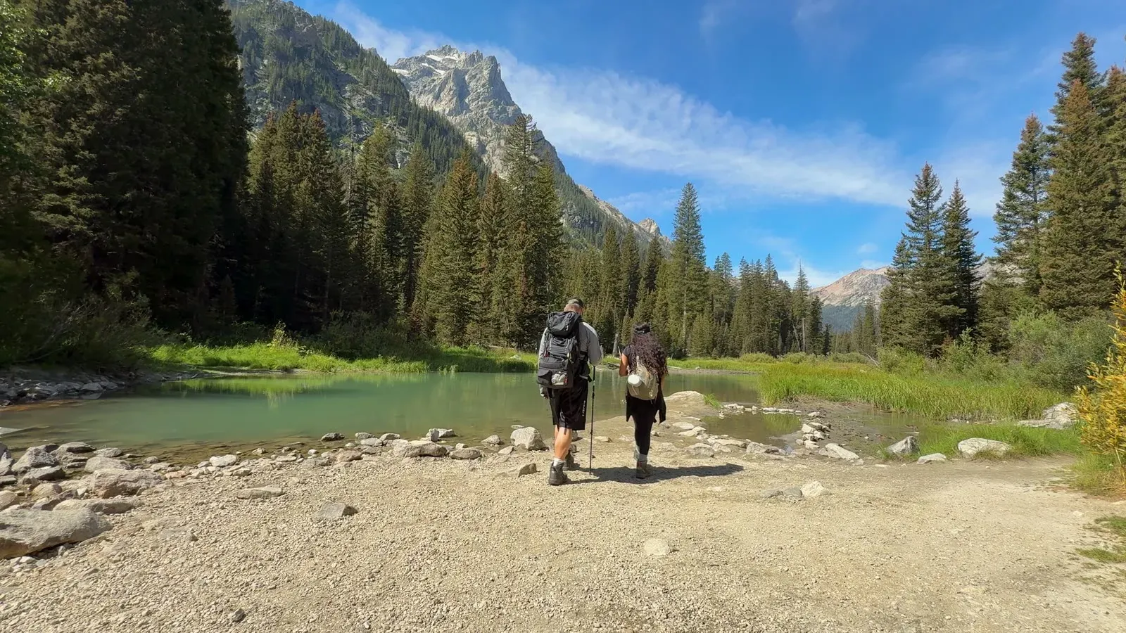 Nathan and Fabiola hiking past a green pond in Cascade Canyon with pine trees and a mountain peak ahead