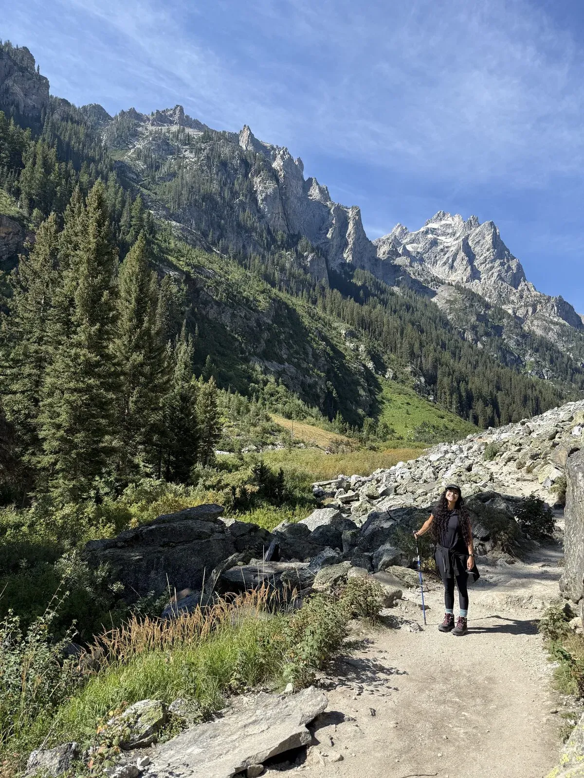 Fabiola with a trekking pole on the rocky Cascade Canyon trail, jagged Teton peaks and blue sky above