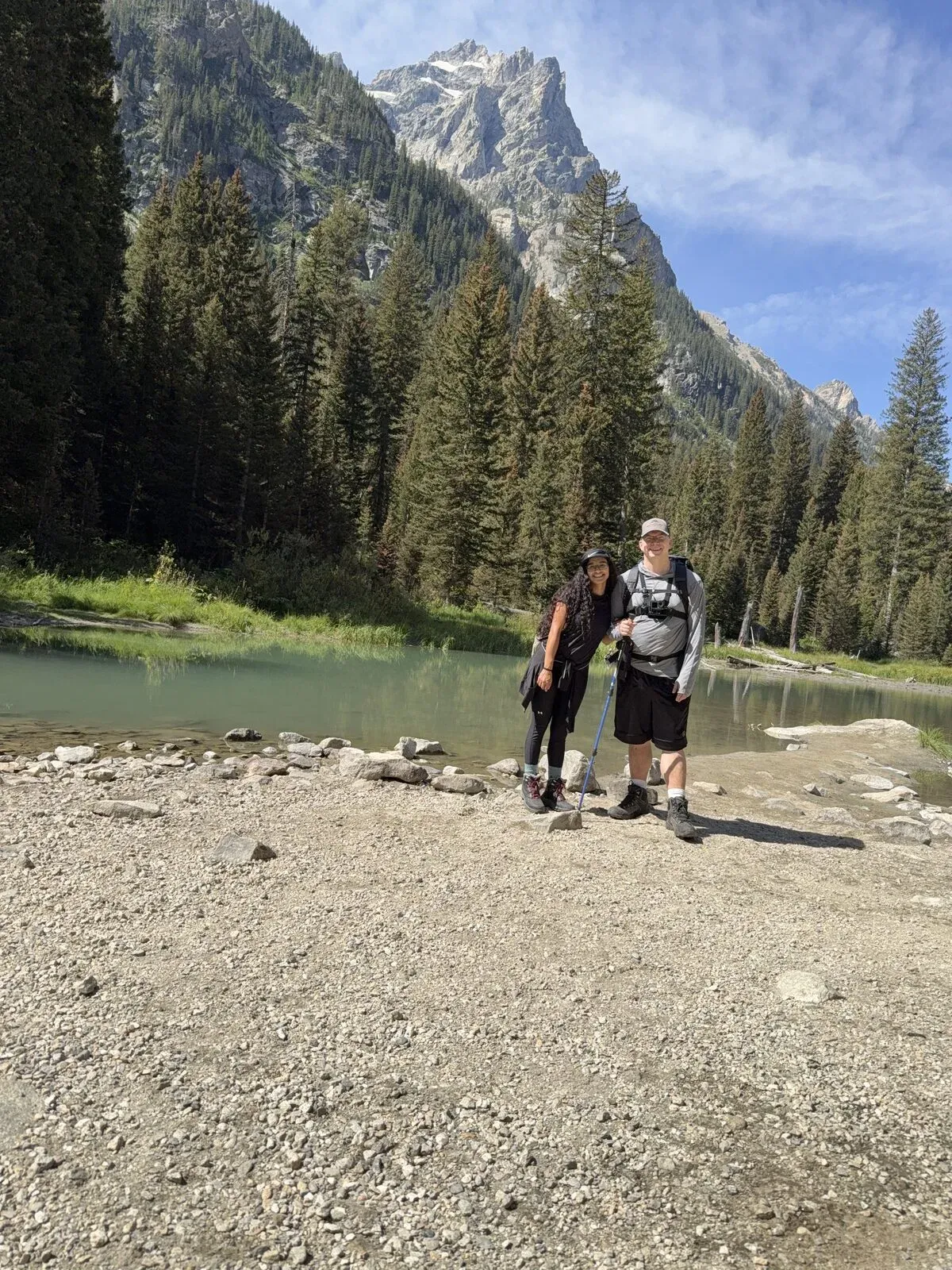 Nathan and Fabiola with trekking poles beside a green pond in Cascade Canyon, pine trees and a Teton peak behind them