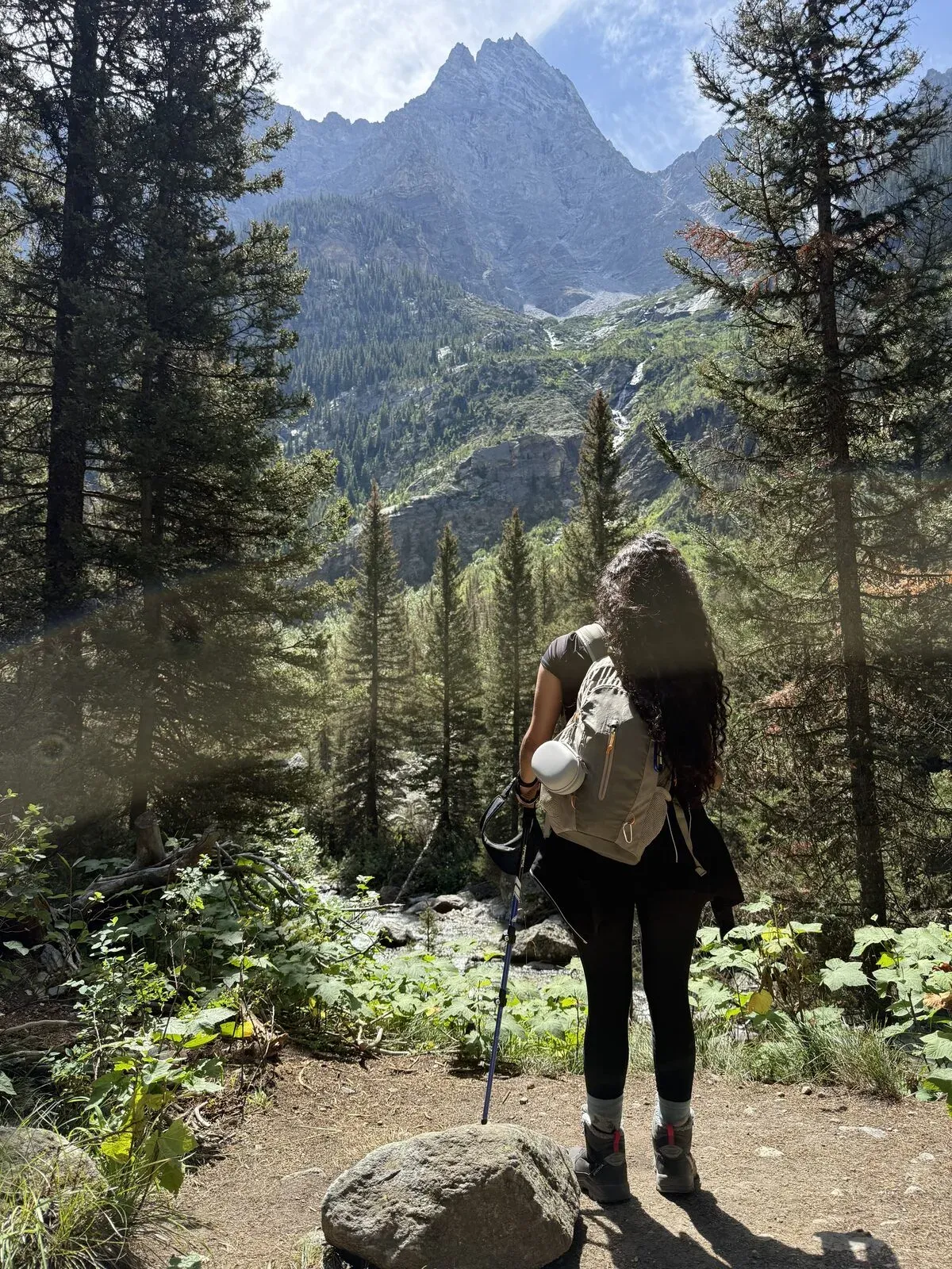 Fabiola from behind with her backpack, looking at a Teton peak through tall pine trees