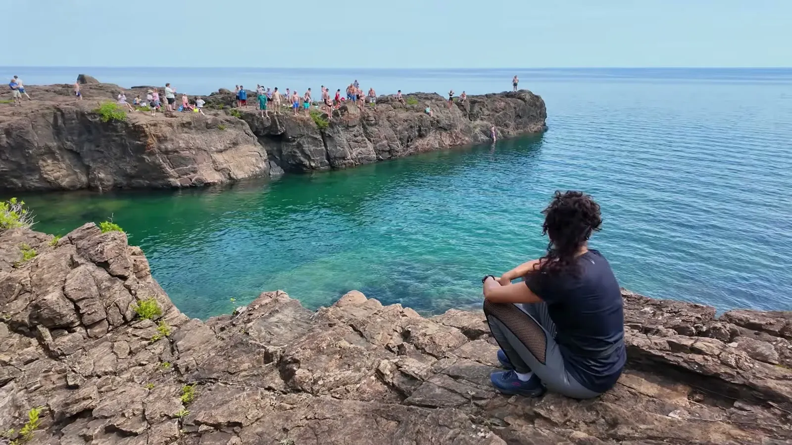 Fabiola sitting on the rocks at Black Rocks, watching a crowd of cliff jumpers lined up above the clear water of Lake Superior.