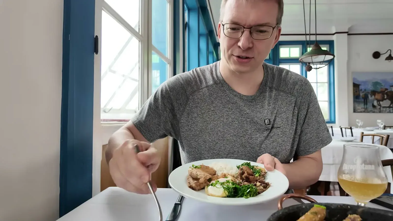 Nathan lifting a plate of feijao tropeiro at Bene da Flauta, with the blue-trimmed dining room and a glass of beer on the table.