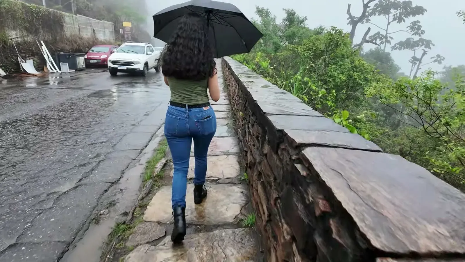 Fabiola walking uphill under an umbrella beside an old stone wall, the street ahead swallowed by fog on the climb to Palacio D'Ouro.