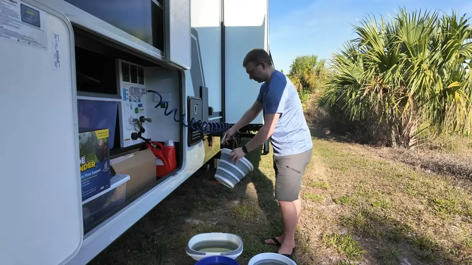 Nathan catches water from the fifth wheel's low-point drain in a collapsible bucket, with more buckets lined up on the grass.