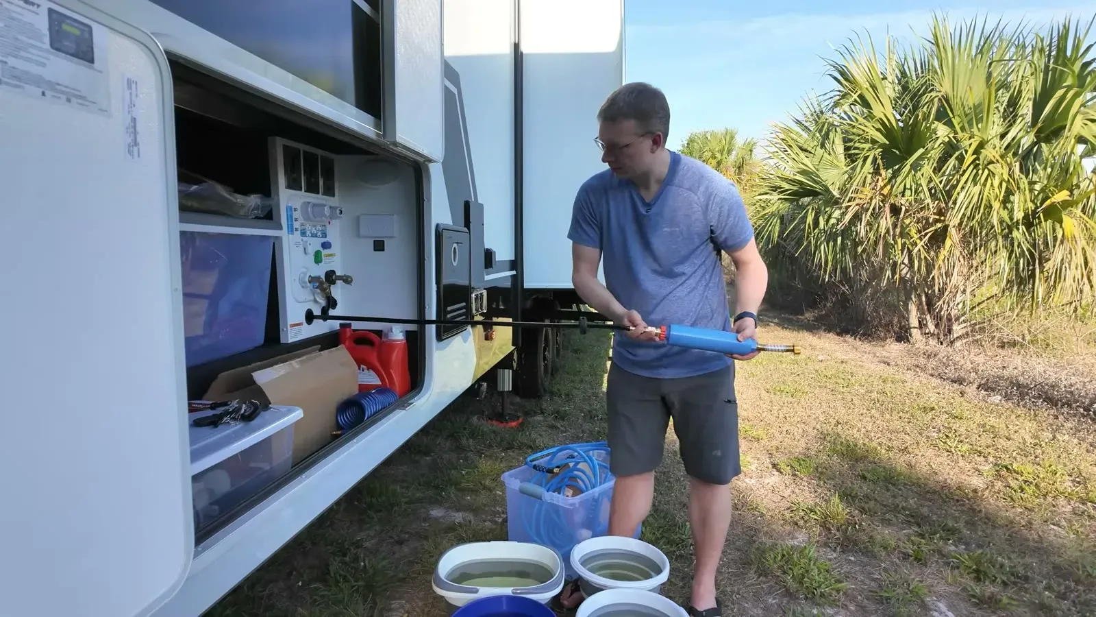 Nathan holds a blue siphon pump over a row of water-filled buckets at the RV's utility bay, ready to send the water back into the tank.