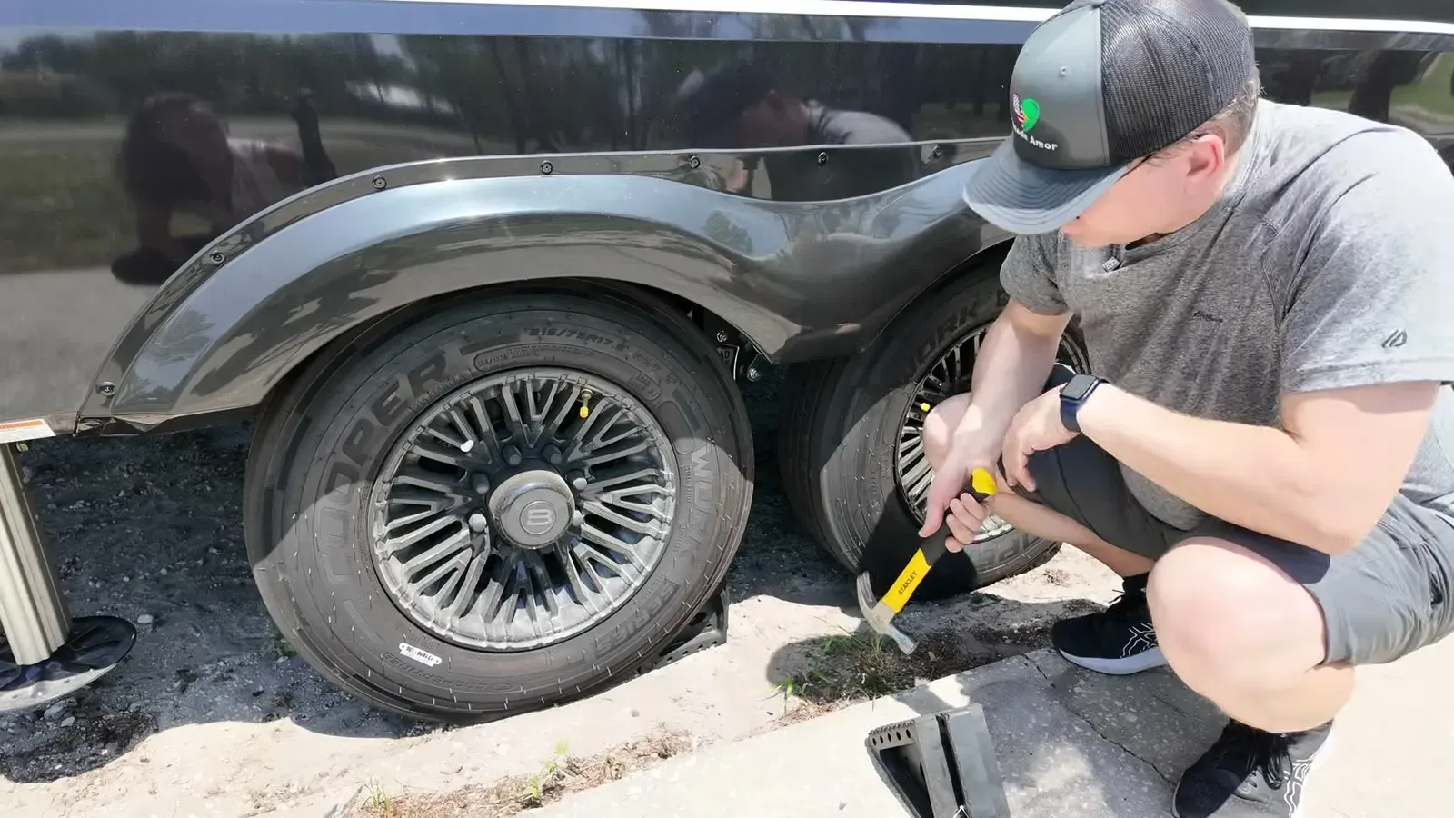 Nathan crouches beside the trailer tires with a hammer, working a wheel chock loose after two months of sitting in one spot.