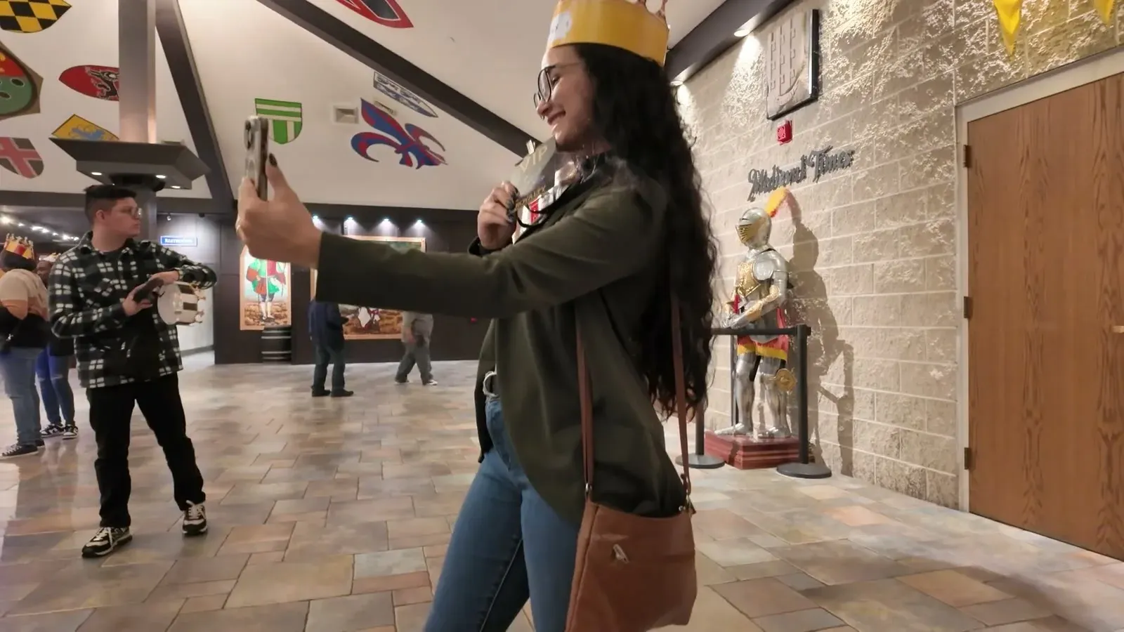 Fabiola snaps a selfie in her paper crown in the castle lobby, with heraldic shields covering the ceiling and a suit of armor by the door.