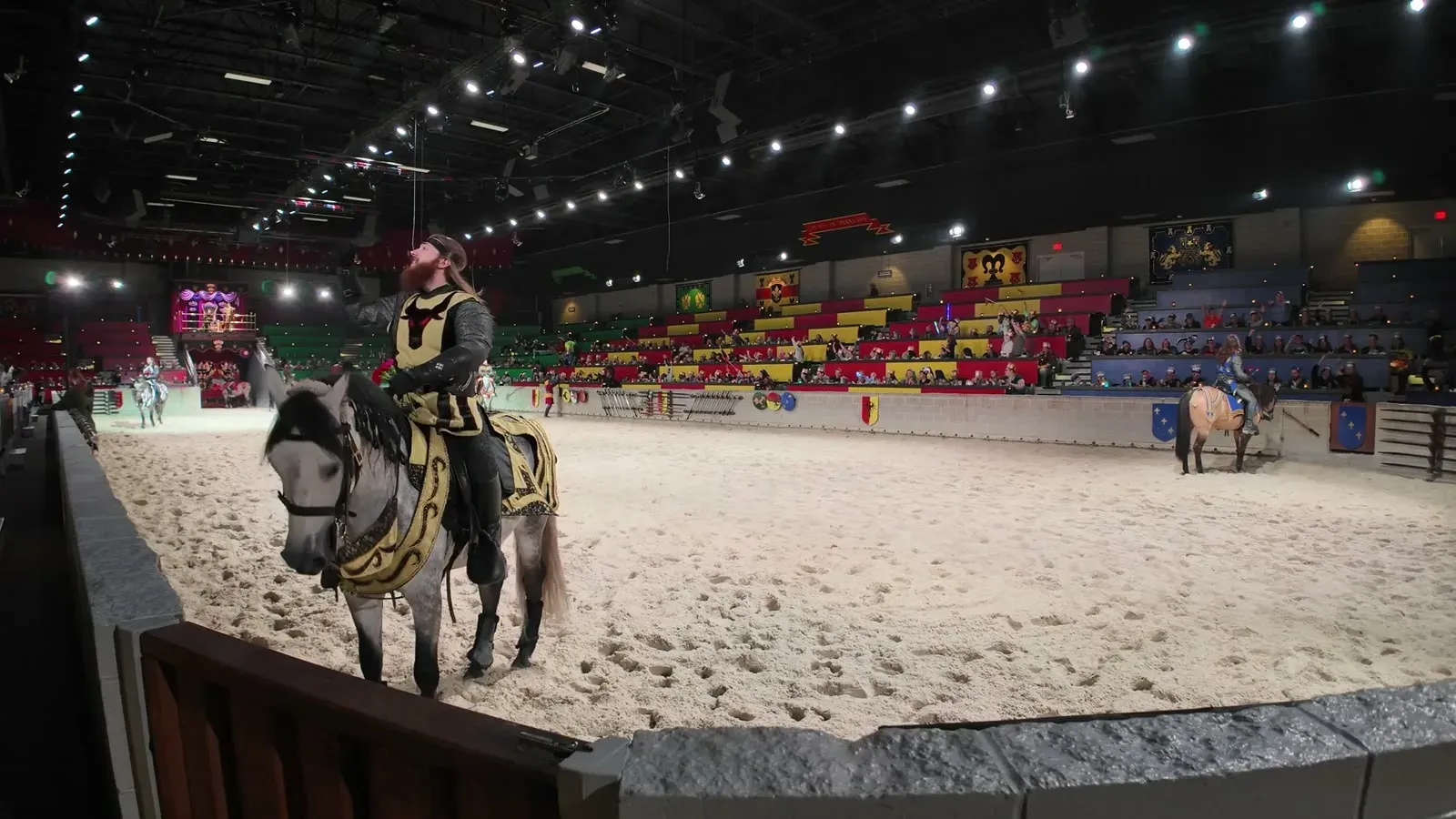A knight in yellow and black rides his horse across the dirt arena floor under the stadium lights.