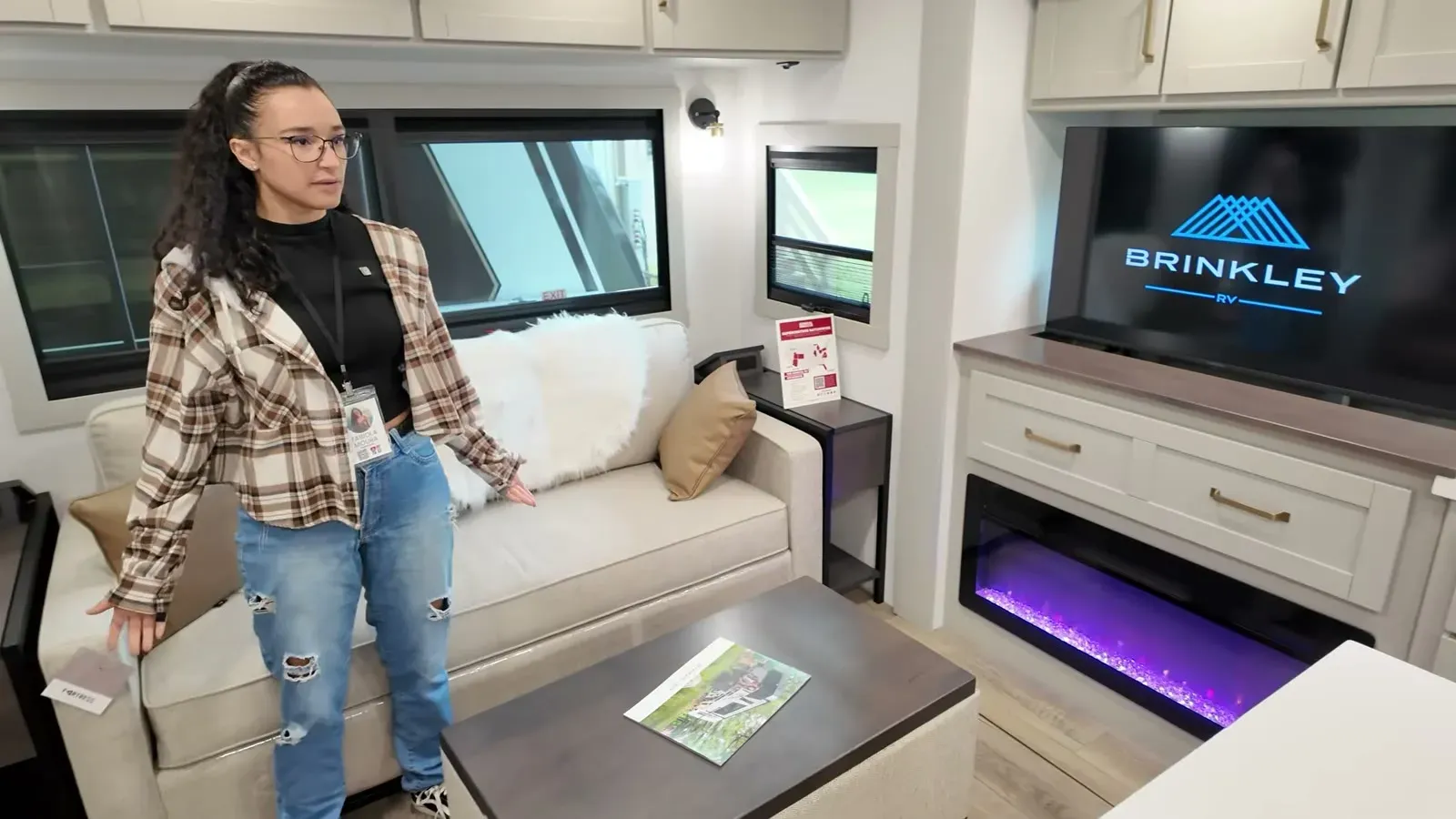 Fabiola showing off the Model Z Air 297 living room, with the sofa, fireplace, and the Brinkley logo up on the TV.