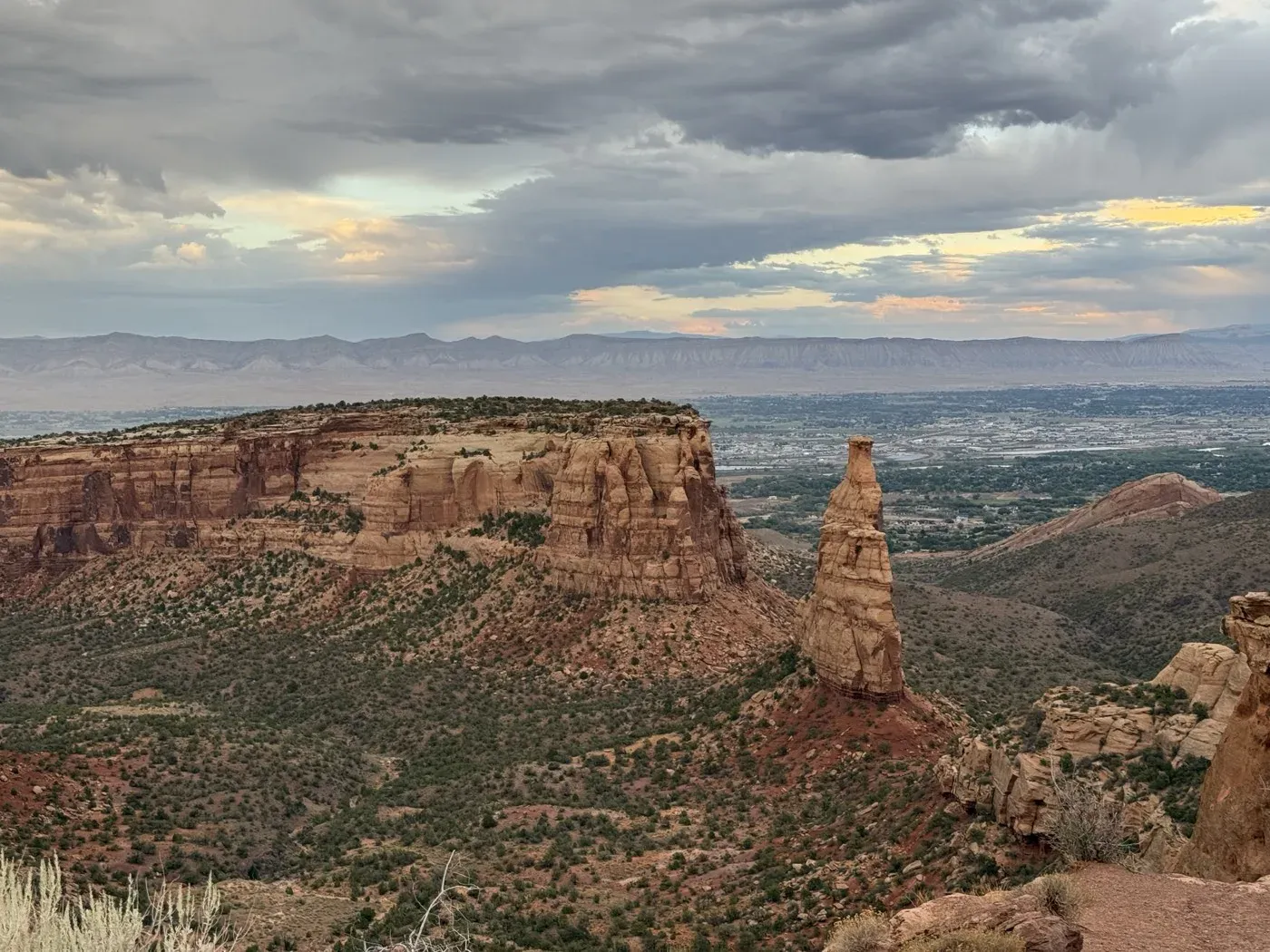 Independence Monument, a lone sandstone tower rising from the canyon at Colorado National Monument, with Grand Junction and a long line of cliffs under storm clouds behind it