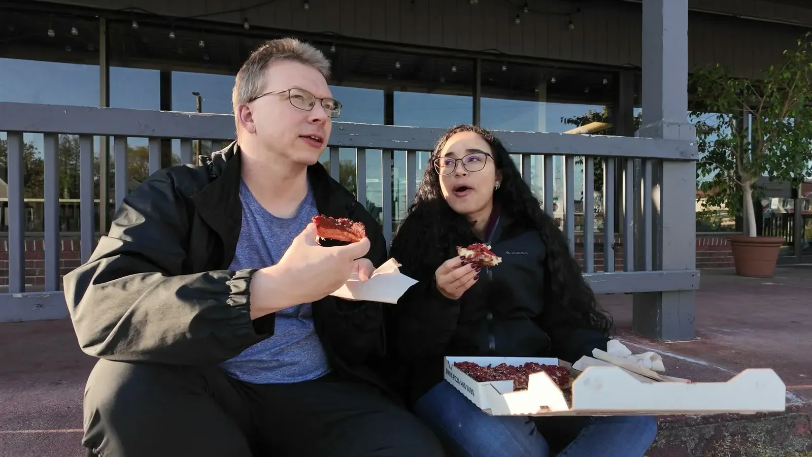 Nathan and Fabiola sharing a Dino's pizza on a bench outside, finally fed and back to talking.