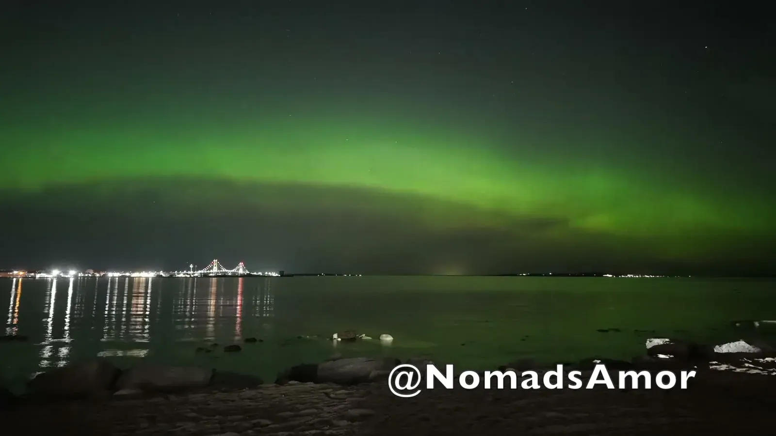 A bright green aurora band arcing over the water while the lit-up Mackinac Bridge glows on the horizon.