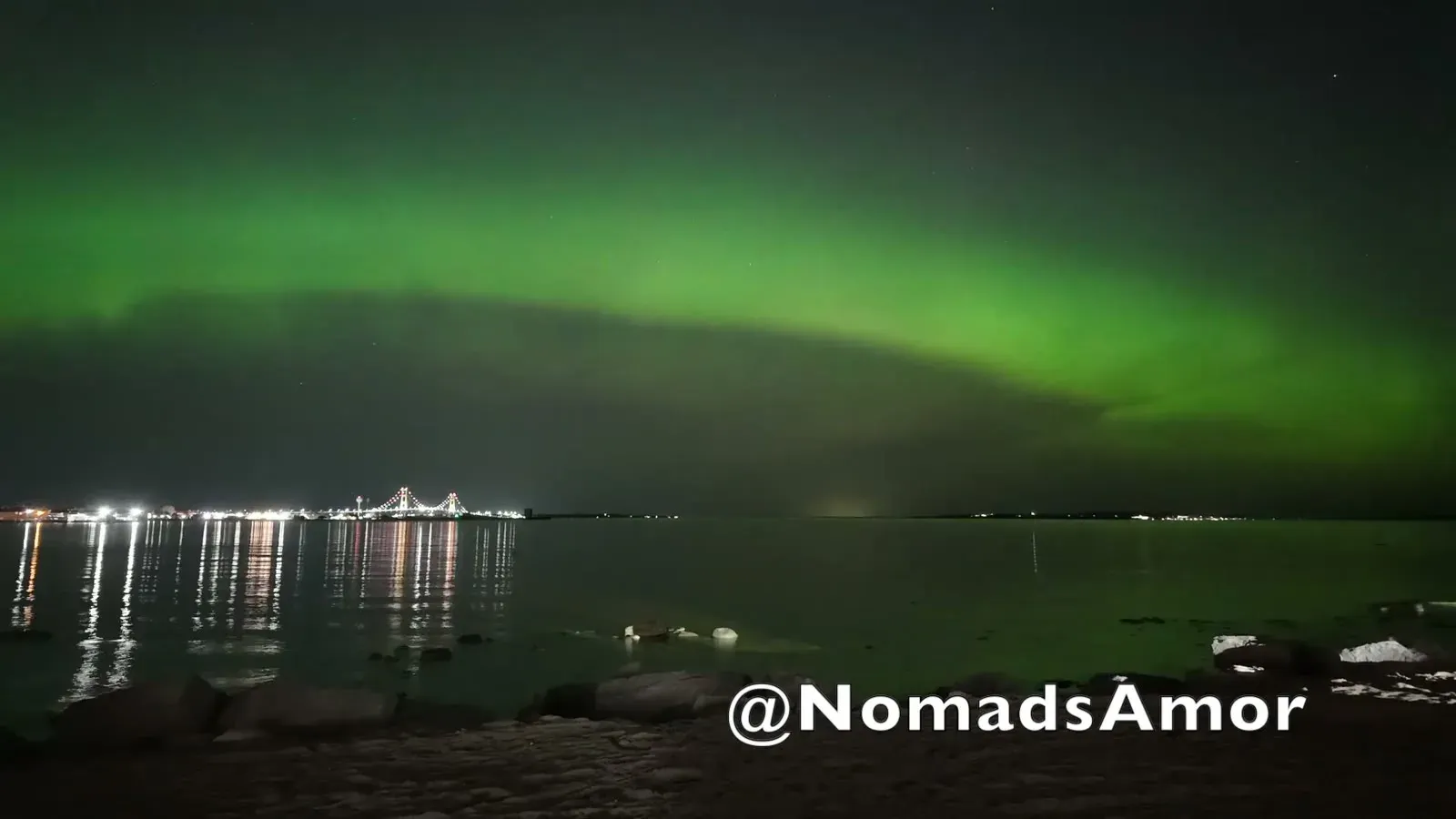The aurora at full strength, a sweeping green arc filling the sky above the Straits with the bridge tiny beneath it.