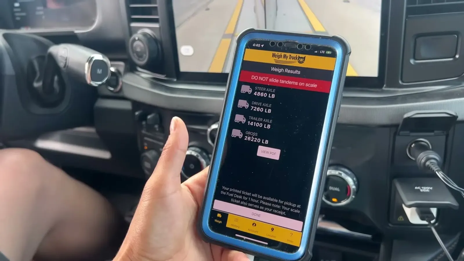 Hand holding a phone in the truck cab with the Weigh My Truck app displaying steer, drive, and trailer axle weights and gross weight