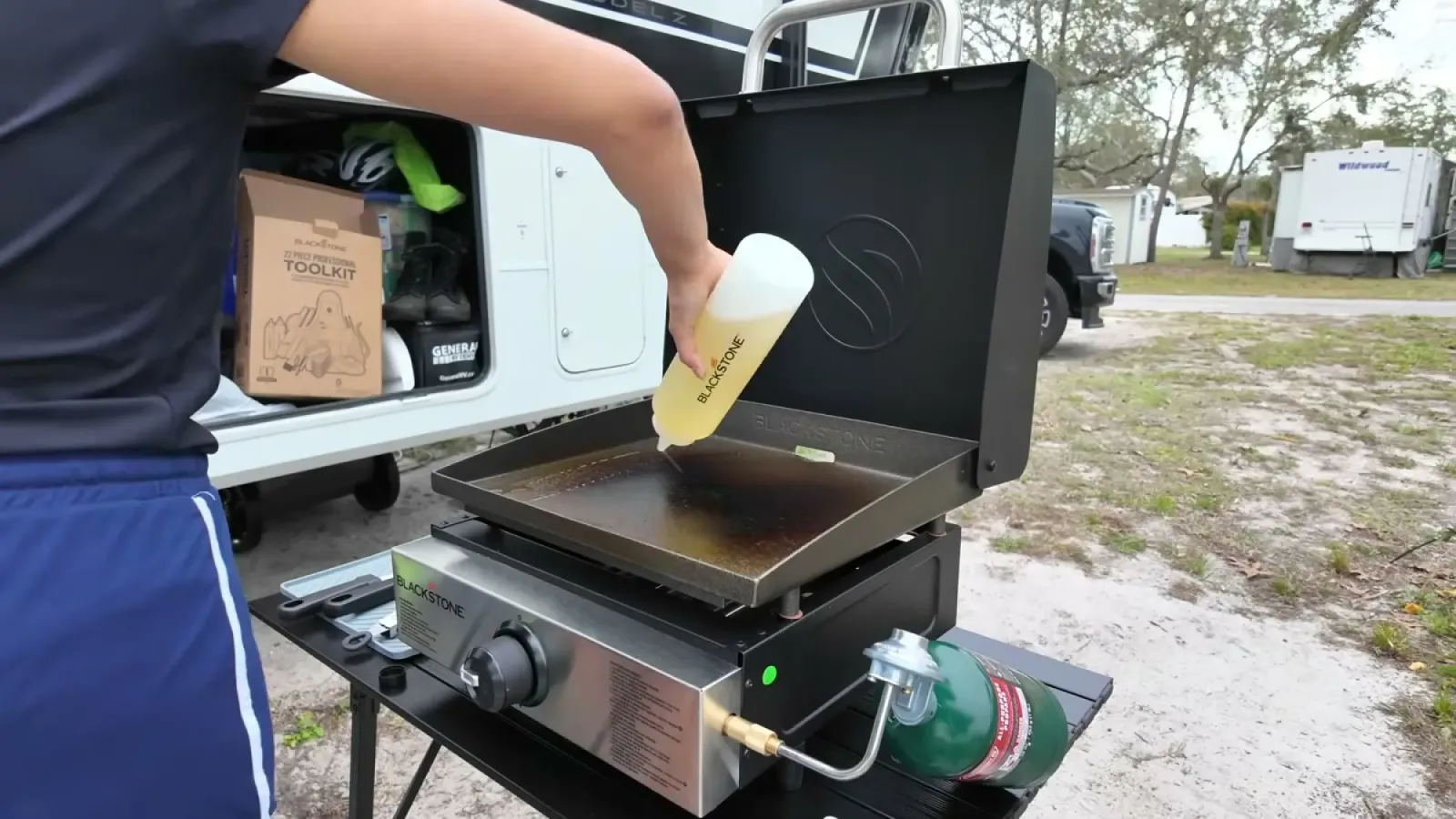 Squeeze bottle pouring oil onto the darkened hot cooktop of the Blackstone griddle during a seasoning round