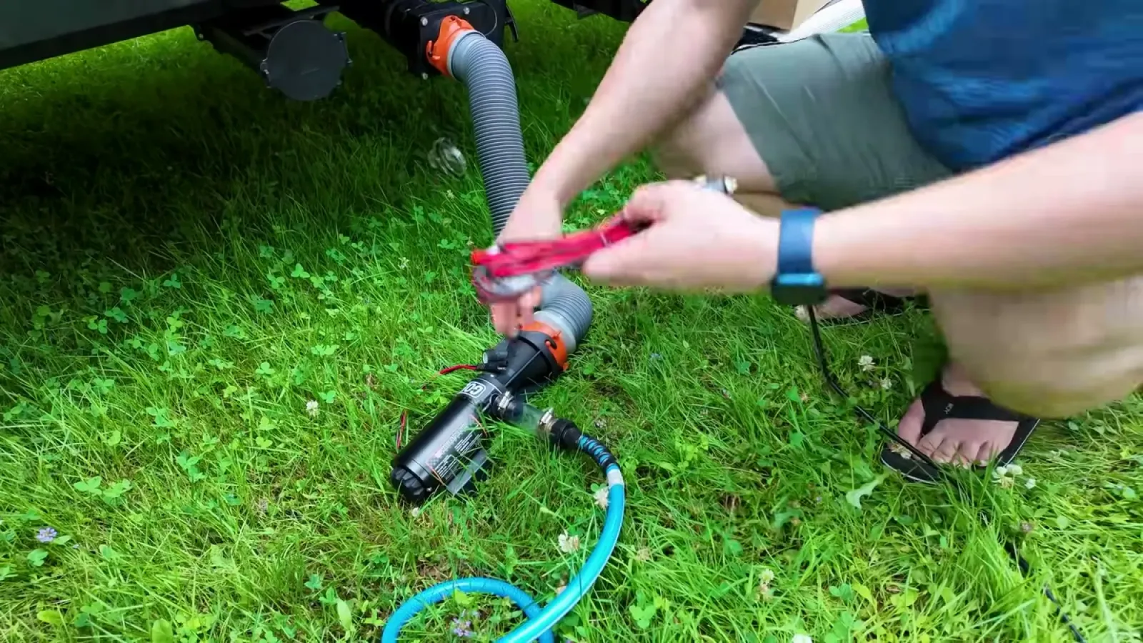 Hands using pliers to clamp the black macerator pump onto the RV sewer outlet hose in the grass, with a garden hose attached