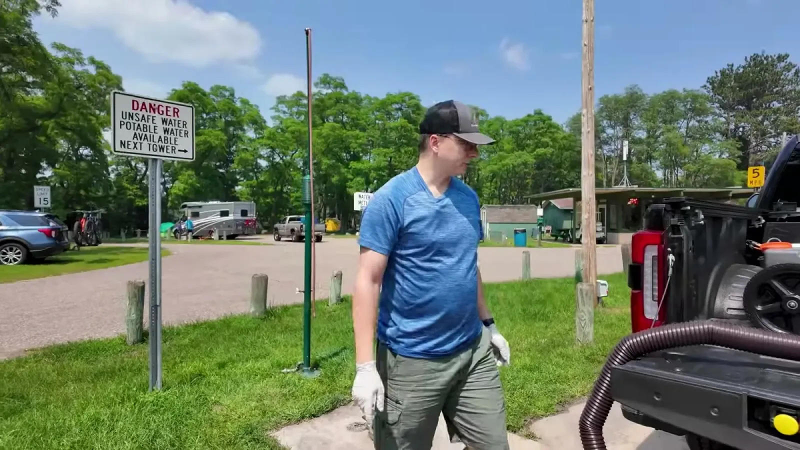 Nathan in gloves walking to the truck at the campground dump station, sewer hose hanging from the tailgate and an unsafe water warning sign nearby