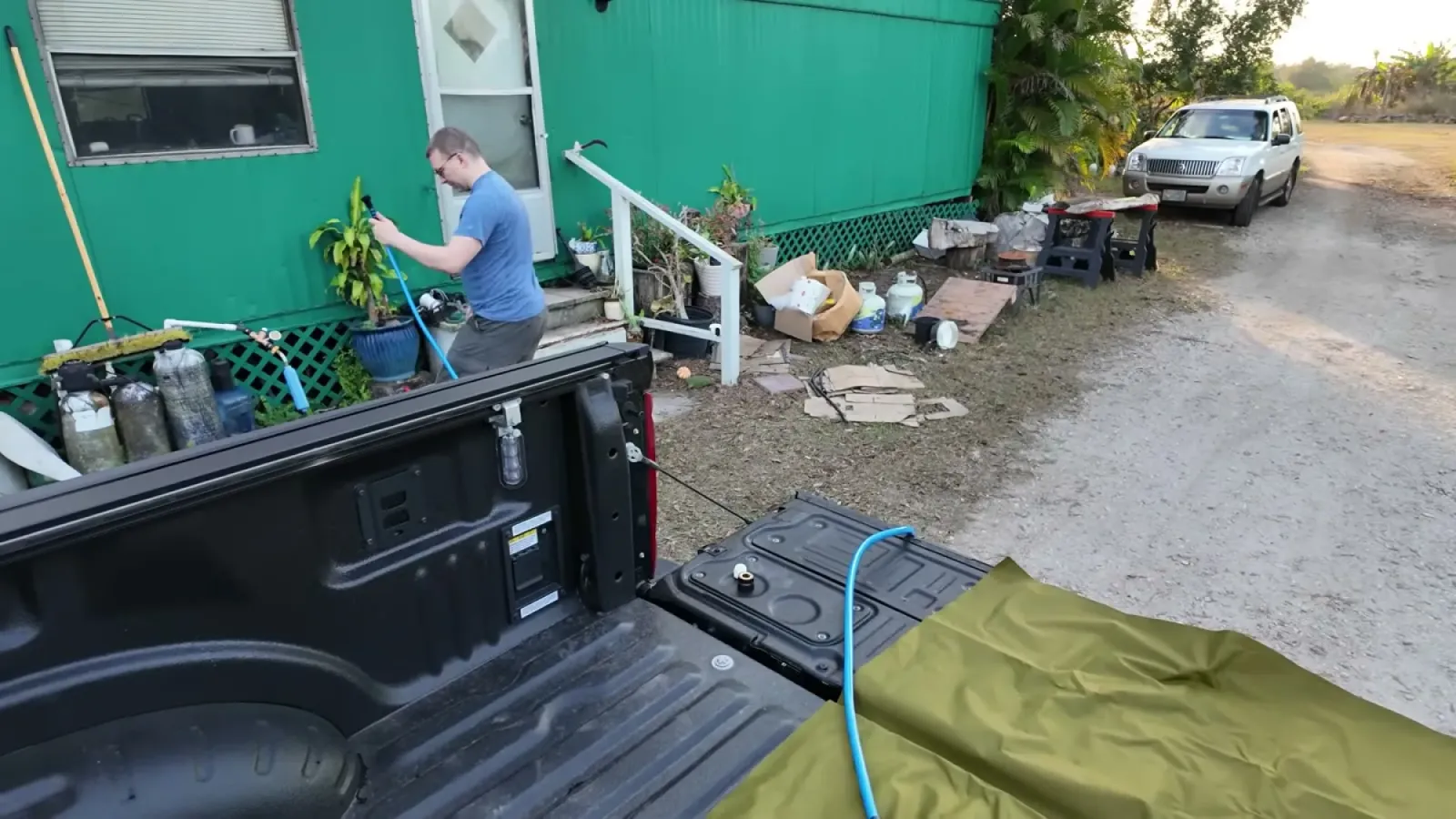 Nathan connecting a blue drinking water hose to a spigot beside the green farmhouse, with the empty water bladder laid out in the truck bed in the foreground