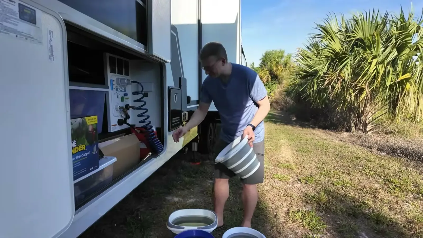 Nathan holding a collapsible bucket at the RV utility bay with more buckets of water on the ground during the fresh tank drain test
