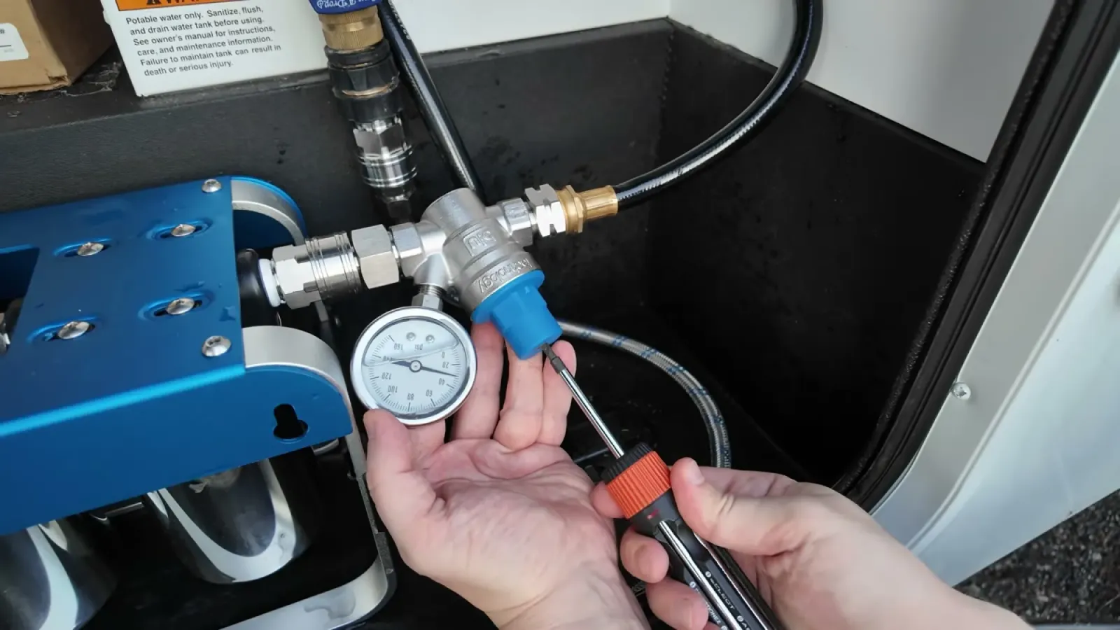 Hand holding a flathead screwdriver next to a water pressure regulator with a round gauge mounted beside the blue filter unit