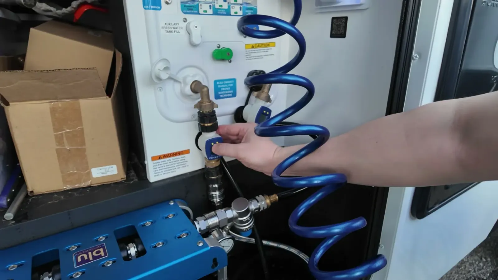 Hand connecting a fitting at the RV water bay panel with a coiled blue hose and the AR3 filter unit below during the first flush