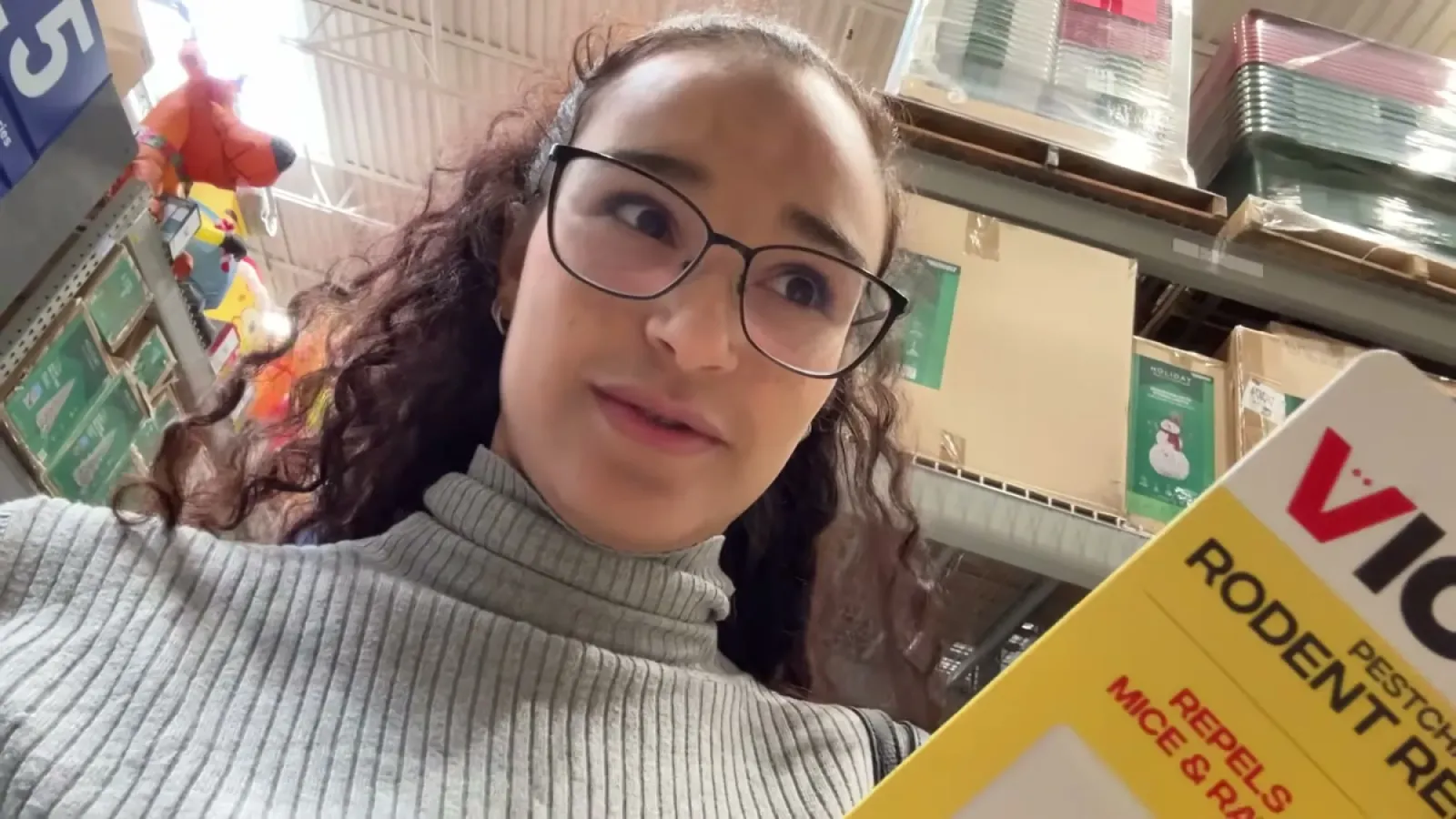 Fabiola in a hardware store aisle holding a boxed rodent repellent product in front of stocked shelves
