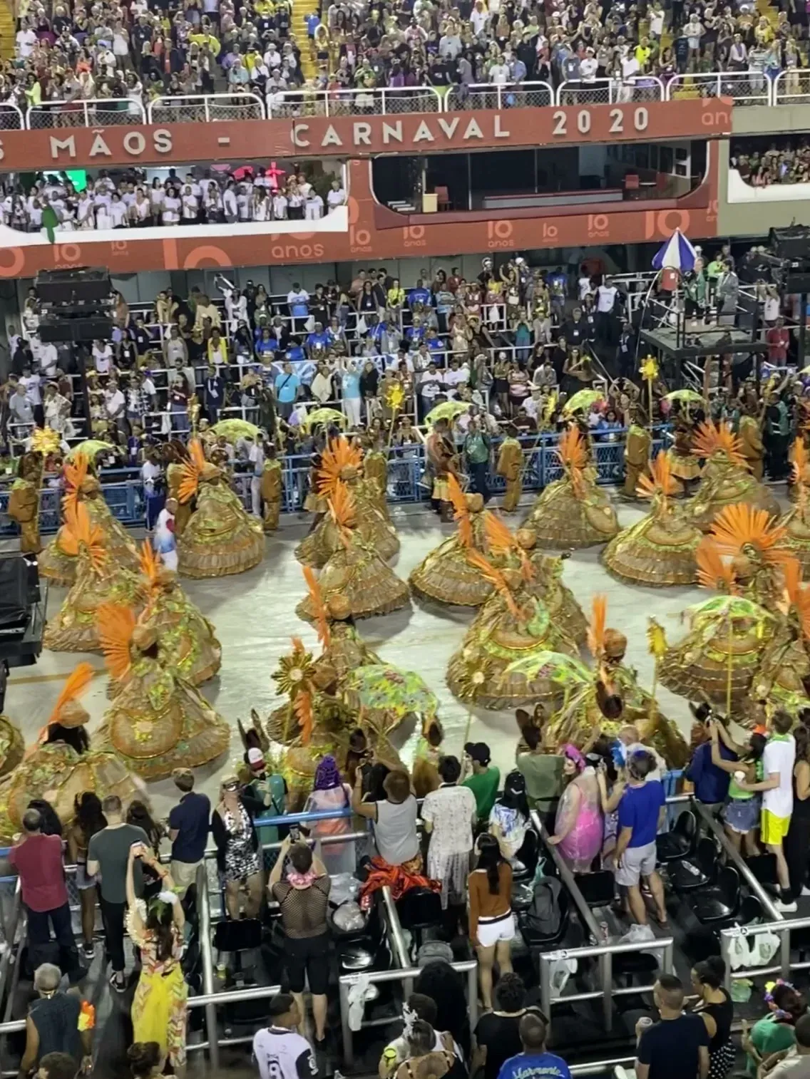The Sambadrome during Carnival 2020 in Rio, packed stands and a parade of gold and orange costumes below a banner reading CARNAVAL 2020.