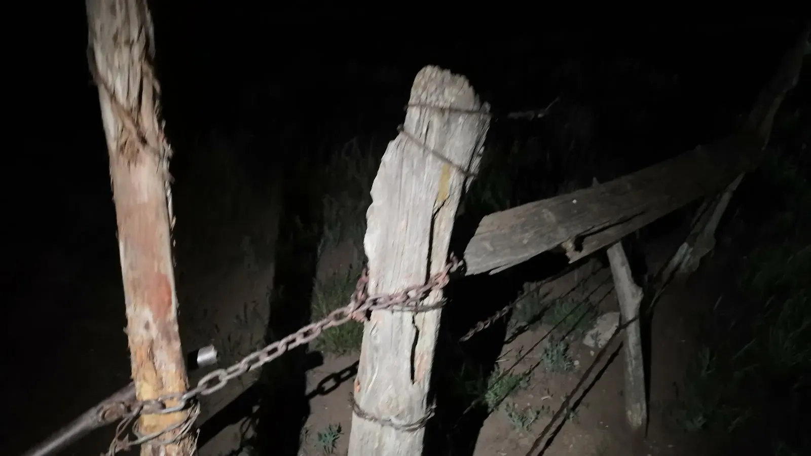 Wooden gate posts with a chain and barbed wire, lit by a flashlight at the grassland entrance at night