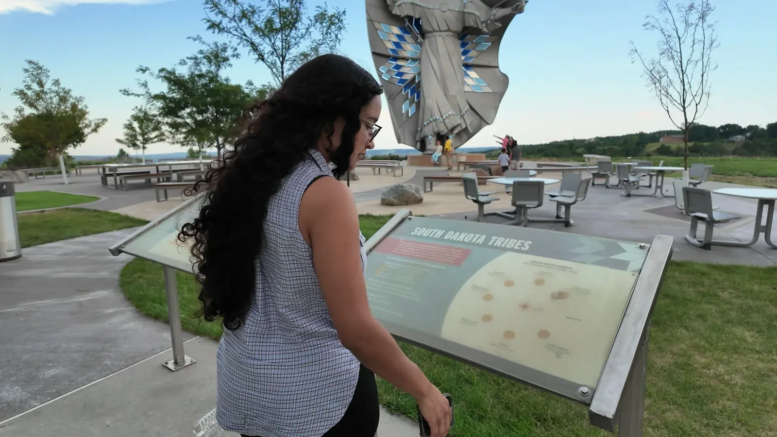 Fabiola reading a South Dakota Tribes information sign at the welcome center, with picnic tables and a tipi shaped shelter behind her