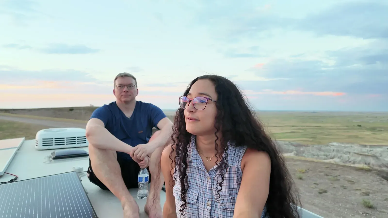 Nathan and Fabiola sitting together on the roof of the fifth wheel at sunset, with the Badlands and open prairie behind them