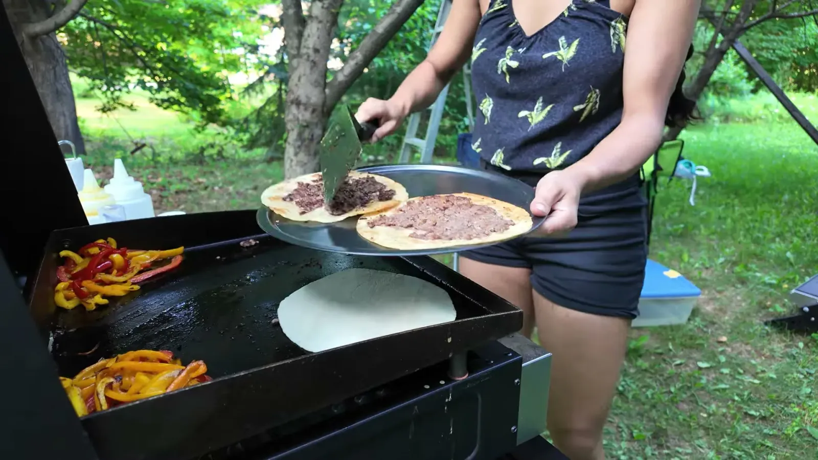Fabiola lowering a raw beef-topped tortilla beef-side down onto the Blackstone griddle, with red and yellow pepper strips cooking in the corners