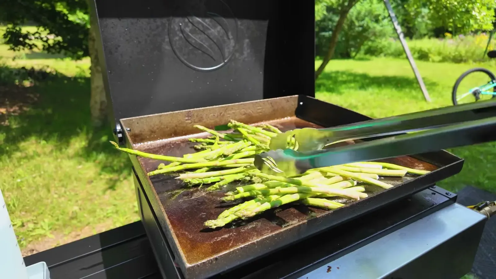 Asparagus spears spread across the Blackstone griddle with tongs, cooking outside the fifth wheel on a summer day