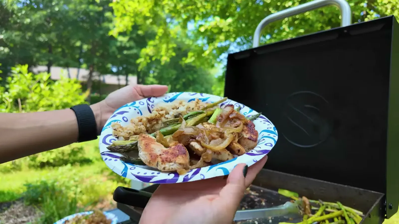 A paper plate of griddled chicken, caramelized onions, asparagus and rice held up in front of the Blackstone